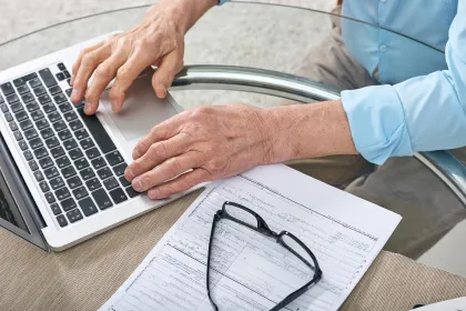 Hände eines Mannes tippen auf einem Laptop neben einem ausgefüllten Formular und einer Brille auf einem Glastisch.