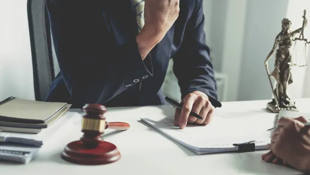 Ein Anwalt im Anzug diskutiert über Dokumente am Schreibtisch, auf dem Bücher, ein Richterhammer, ein Siegel und eine Statue der Justitia liegen.