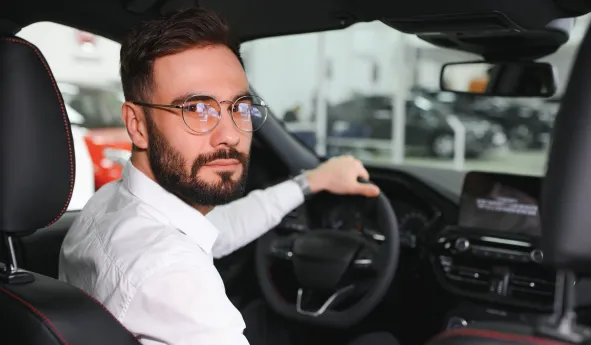 Ein junger Mann mit Brille sitzt am Steuer eines Autos und schaut in die Kamera.