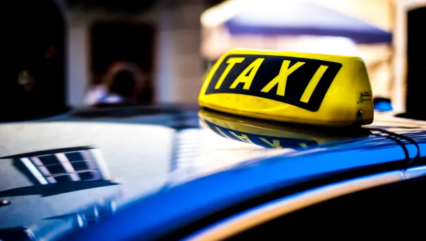 Gelbes Schild mit der Aufschrift "Taxi" auf dem Dach eines Autos vor einer Stadtstraße.