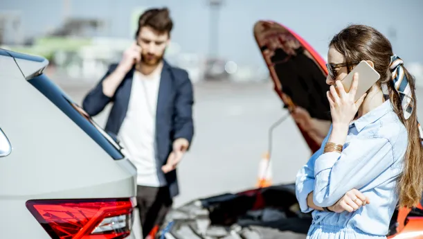 Ein Mann und eine Frau telefonieren neben einem Auto mit geöffneter Motorhaube nach einem Unfall.